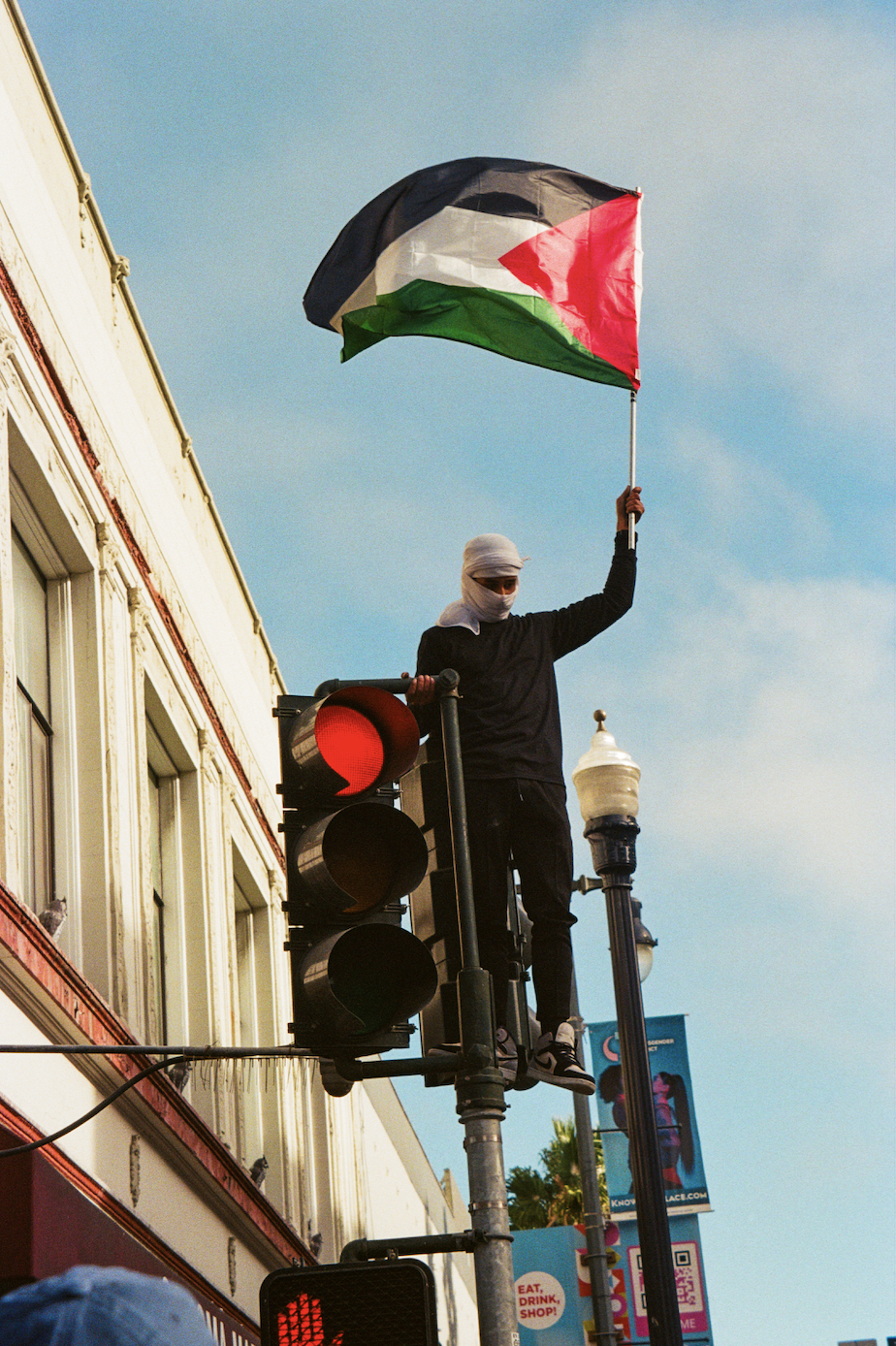 Protestor in a white face covering waves a Palestinian flag. This person stands on top of a traffic light, leaning on the light itself which comes up to his chest. He looks down at protesters (not pictured) and behind him are brick red and white buildings and banner advertisements on poles lining the street behind him.